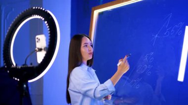 Canlı yayında öğretmenlik. Eğitmen, daha iyi görebilmek için halka ışığını kullanırken cam bir tahtaya yazı yazarak kavramları gösteriyor.