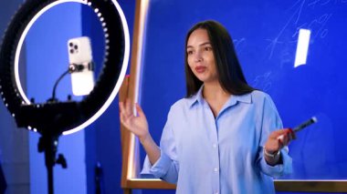 Kameradaki genç kadın. Modern bir stüdyoda, genç bir kadın elinde kalem tutarken izleyiciyle iletişim kurar, kendinden emin ve hareketli görünür..