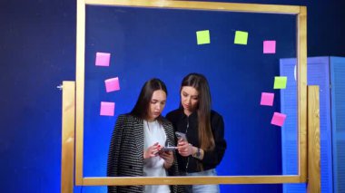 Uzayda telefon kullanan arkadaşlar. İki genç kadın, modern bir çalışma alanında renkli yapışkan notlarla çevrili akıllı telefonlarıyla meşgul oluyor..