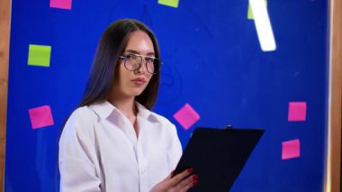 Yaratıcı bir toplantıya liderlik eden genç bir kadın. Gözlüklü genç bir kadın, yapışkan notlarla dolu canlı bir toplantı yerinde bir pano tutuyor..