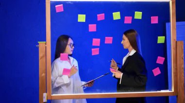 Yapışkan notlarla kadın takım çalışması. İki kadın beyin fırtınası sırasında renkli yapışkan notlarla kaplı cam bir tahtanın önünde fikirleri tartışıyor..