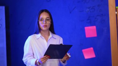 Genç yetişkin toplantı için hazırlanıyor. Elinde bir pano tutan genç bir kadın mavi ışıklı bir odada duruyor. Etrafı notlar ve çizelgelerle çevrili, tartışmaya hazırlanıyor..
