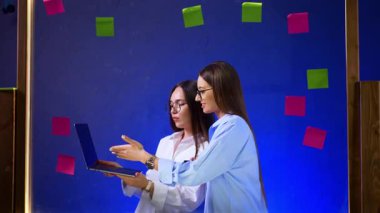 Dizüstü bilgisayarlarda birlikte çalışan kadınlar. İki kadın, mavi arka planda renkli yapışkan notlarla çevrili bir dizüstü bilgisayarla çalışırken fikirleri tartışıyor..