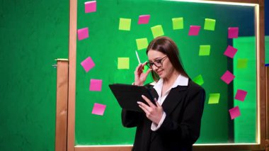 Beyin fırtınasında yaratıcı kadın. Siyah takım elbiseli genç bir kadın yeşil arka planda renkli yapışkan notlarla çevrili bir tablete yazıyor..
