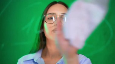 Genç bayan yeşil tahtaya yazıyor. Mavi tişörtlü ve gözlüklü bir kadın, canlı yeşil bir tahtaya bir kalemle yaratıcı bir faaliyet sergiliyor..