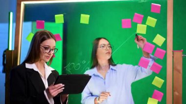 Yaratıcı bir çalışma alanında işbirliği. İki kadın, dijital tablet ve fikir üretmek için yapışkan notlar kullanarak modern bir ofiste iş birliği yapıyor..