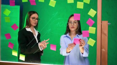 Yapışkan notlarla beyin fırtınası yapan profesyoneller. Modern bir çalışma alanında renkli yapışkan notlarla kaplı cam bir yüzey üzerine beyin fırtınası yapan iki kadın..