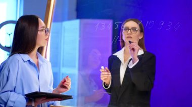 Profesyoneller birlikte modern bir şekilde çalışıyorlar. Çağdaş bir ofis ortamında cam tahtaya yazı yazarken, iki profesyonel bir tartışma içinde..