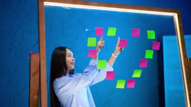 Kadın notlarla kalp yaratır. Genç bir kadın neşeli bir şekilde parlak bir odada cam bir yüzey üzerinde kalp şeklinde renkli yapışkan notlar düzenler..