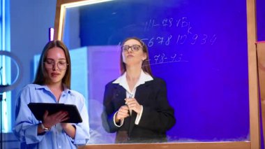 Genç kadınların olduğu bir sınıf. İki genç kadın bir sınıf tartışmasına giriyorlar. Biri tablet tutarken, diğeri şeffaf bir levhada açıklıyor..