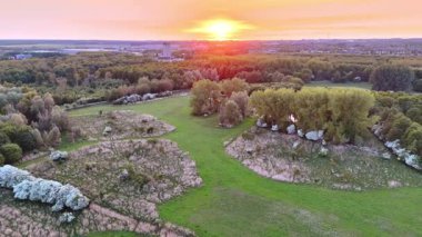 Yeşil ormanlarla çevrili çayırların üzerinde uçuyorlar. Arkaplanda pembe gökyüzünde batan güneş.