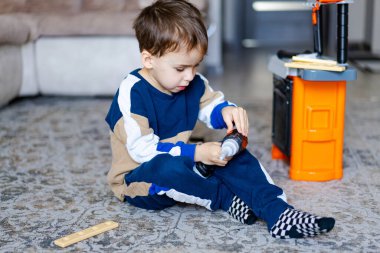 Çocuk oyuncak aletlerle oynuyor. Oyuncak matkap kullanmaya odaklanmış, oyun aletleri ve parlak bir tezgahla çevrili genç bir çocuk yerde oturuyor..
