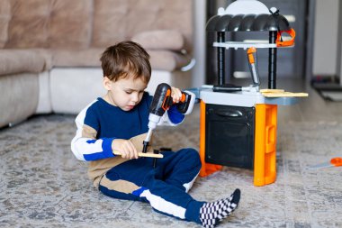 Çocuk içeride oyuncak aletlerle oynuyor. Genç bir çocuk yerde oturuyor, oyuncak matkapla rahat bir oturma odasında mutfak seti yapıyor..