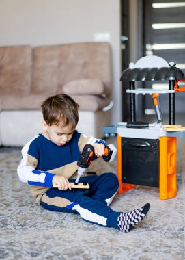 Çocuk oyuncak aletlerle oynuyor. Genç bir çocuk oturma odasının zeminindeki mutfak setini tamir etmek için oyuncak matkap kullanıyor..