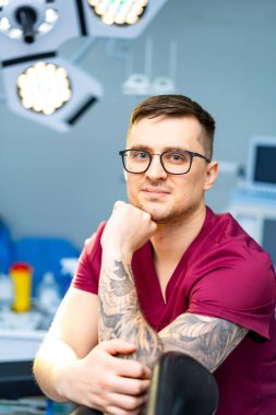 Önlüklü doktor ofisinde. Kendine güvenen bir tıp uzmanı klinikte oturur, dövme yaptırır ve gözlük takar, hastalara yardım etmeye hazırdır..