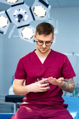 Cerrah ameliyathanede aletleri ayarlar. Kestane rengi önlüklü bir cerrah, iyi aydınlatılmış bir ameliyathanede otururken cerrahi aletleri inceliyor..