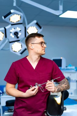 Parlak operasyon için cerrah hazırlıkları. Derin bordo önlüklü bir cerrah iyi aydınlatılmış bir ameliyathanede dururken cerrahi aletler tutar..