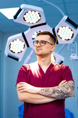 Ameliyat önlüklü, dövmeli bir cerrah. Bir cerrah, kolları çaprazlanmış, önlük takmış ve parlak cerrahi ışıkların altında dövmelerini sergiliyor..