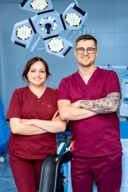 Modern bir klinikteki doktorlar. İki sağlık çalışanı, iyi aydınlatılmış bir klinikte birlikte duruyorlar. Hasta bakımına hazırlar, önlük giyer ve dostça gülümserler..