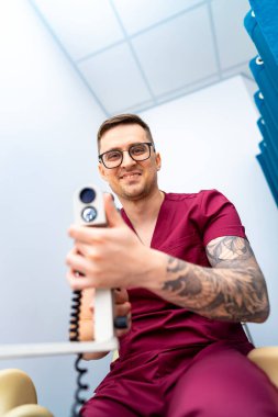 Tıbbi aletler kullanan bir klinisyen. Bir sağlık çalışanı elinde tıbbi bir cihaz tutarken gülümseyerek klinik ortamda dostane bir atmosfer sergiliyor..