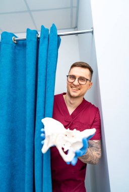 Doktor tıbbi model gösteriyor. Bir sağlık uzmanı, klinik bir ortamda mavi bir perdenin arkasında dururken bir iskelet modeli tutar..