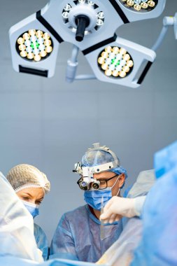 Cerrahi ekip operasyon halinde. Tıp uzmanları steril bir ortamda birlikte çalışıyorlar. Parlak ışıklı bir cerrahi müdahaleye odaklanıyorlar..