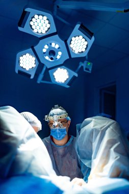 Cerrahlar parlak odada ameliyat yaparlar. İki cerrah, modern bir ameliyathanenin ışıklarıyla aydınlatılmış bir cerrahi müdahalede bulunuyorlar..