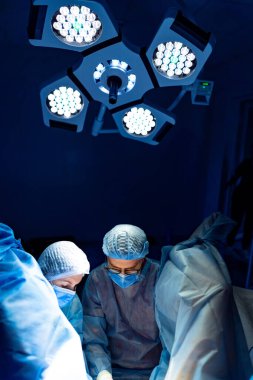 Ameliyathanede cerrahlar var. Ameliyat önlüğü ve maskeli iki cerrah steril bir ortamda parlak ışıklar altında cerrahi bir prosedüre odaklanıyor..