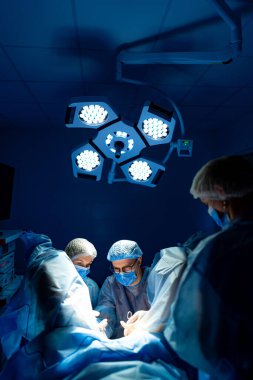 Hastanede çalışan cerrahlar var. Cerrahlardan oluşan bir ekip, hastane ortamındaki parlak ameliyathane ışıkları altında cerrahi bir işleme tabi tutuluyor..