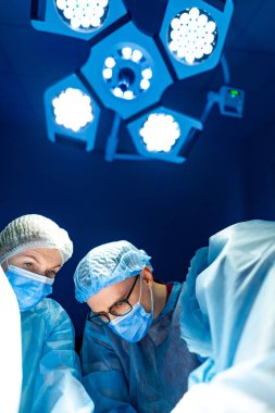 Cerrahlar hastanede ameliyat yapıyor. Tıbbi ekip steril bir ortamda, parlak ışıklar altında hasta bakımına odaklanmış bir ameliyat gerçekleştiriyor..