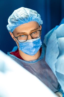 Cerrah ameliyata odaklanmış. Ameliyat önlüğü ve maske takmış bir cerrah hastane ortamında çok önemli bir cerrahi prosedüre hazırlanıyor..