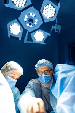 Cerrahlar ışıkların altında çalışıyor. Mavi önlüklü cerrahlar, iyi aydınlatılmış bir ameliyathanede ameliyat sırasında bir hastaya odaklanırlar..