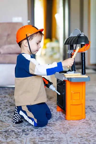 Oyuncak mutfak setiyle yemek yapan genç bir çocuk. Genç bir çocuk, oyuncak mutfak seti ve ahşap çatal bıçaklarla oynarken bir kask takarak halıya diz çöker..