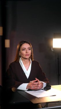 Odaklanmış kadın muhabir masada kamerayla konuşuyor. Stüdyoda blog görüntüleri. Arka planda ışık. Dikey video.
