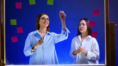 Cam tahtanın arkasında gözlüklü güzel, pozitif esmerler var. Kadın tahtaya siyah kalemle bir grafik çiziyor..