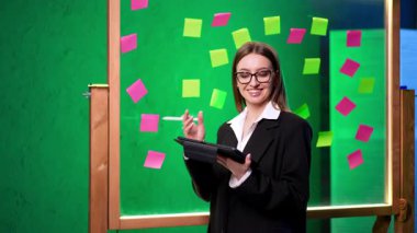 Gözlüklü, siyah ceketli ve beyaz gömlekli güzel bir bayan cam tahtanın yanında duruyor. Kadın önce biriyle konuşur, sonra tabletine bakar..