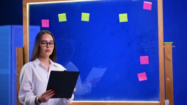 Beyaz gömlekli ve gözlüklü beyaz bir kadın elinde bir dosya tutuyor. Hanımefendi iş sunumu için hazırlanıyor. Arkaplanda çıkartmaları olan cam tahta..