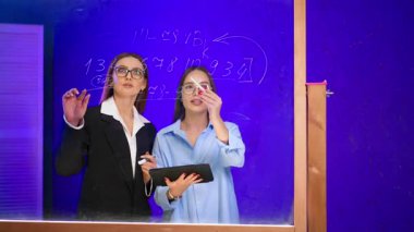 Beyaz kadınlar tablete bakar, iletişim kurar ve cam panodaki rakamları karşılaştırırlar. Siyah ceketli kadın tahtaya yazıyor. İş konsepti.