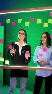 Beyaz kadınlar cam levhanın arkasında durarak iletişim kurarlar. Hanımlar toplantı odasındaki cam duvar planlayıcısında beyin fırtınası yaparken rakamları tartışırlar. Dikey video.
