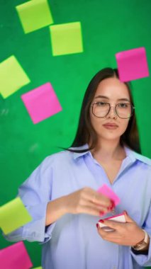 Gözlüklü ve gömlekli beyaz kadın cam tahtaya renkli çıkartmalar yapıştırıyor. Finansal sunum konsepti. Dikey video.