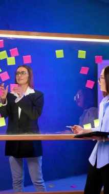 Esmer kadınlar cam tahtanın iki tarafında dururlar. Siyah ceketli kadın çıkartmaları tahtadan indiriyor. Beyin fırtınası konsepti. Dikey video.