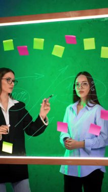 Ofisteki cam panoda iki bayan danışmanlık yapıyor. Kadınlar etiketlere yazılmış rakamları ve fikirleri tartışırlar. Dikey video.