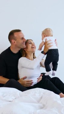 Kafkasyalı ebeveynler yatakta oturup sevgili bebekleriyle oynuyorlar. Anne çocuğunu başının üstünde büyütür ve ebeveynler bir çocuğa gülümser. Dikey video