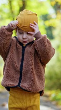 Kahverengi kabarık ceket giyen küçük bir çocuk. Bebek dışarıda sarı şapkasını çıkartıyor. Arkaplan bulanık. Dikey video