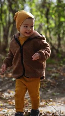 Parkta güzel beyaz bir bebek koşuyor. Sonbahar doğasında yürüyen şapkalı ve ceketli gülümseyen bir çocuk. Dikey video