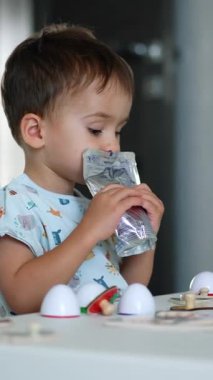 Masada oturan sevimli çocuk doypack 'ten meyve suyu içiyor. Toddler önündeki oyuncaklara odaklanmış görünüyor. Dikey video