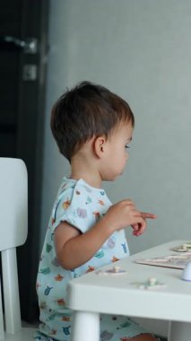 Küçük çocuk önündeki masada duran bulmacalara bakıyor. Sevimli oğlan ellerini heyecanla sallıyor. Dikey video