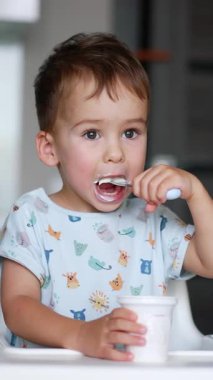 Sevimli çocuk masada oturup plastik bardaktan yoğurt yiyor. Bebek kaşıktan yemek yiyor. Arkaplan bulanık. Dikey video
