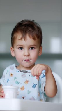 Sakin odaklı erkek bebek yoğurt yiyor. Çocuk bir bardaktan biraz süt ürünü almak için kaşık kullanıyor. Arkaplan bulanık. Dikey video