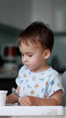 Küçük çocuk plastik bardaktan yoğurt yiyor. Küçük çocuk bir kaşık kullanır ve onu kendisi doldurur. Arkaplan bulanık. Dikey video